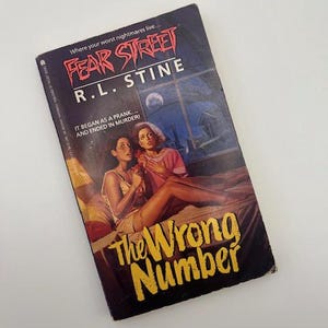 May include: A vintage paperback book titled "Fear Street: The Wrong Number" by R.L. Stine. The cover features two women in a bedroom with a full moon visible through a window. The book's title is in large, yellow, stylized letters.