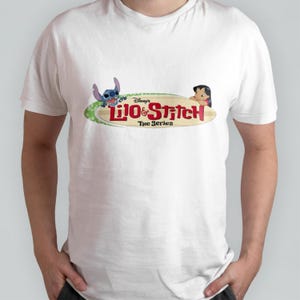 May include: White t-shirt featuring a graphic of Lilo & Stitch: The Series. The graphic depicts Lilo and Stitch on a surfboard with the show's title in red.