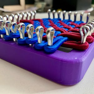 May include: A purple rectangular knitting loom with silver hooks. Blue and red yarn is woven across the loom, creating a textured pattern. The loom is designed for crafting projects.