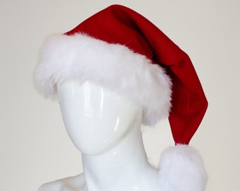 Puede incluir: Un gorro de Papá Noel rojo con un borde de piel sintética blanca. El gorro tiene una punta larga y puntiaguda y un pompón en el extremo.