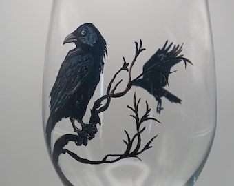 May include: A clear wine glass with a black raven design. The raven is perched on a branch with other branches and leaves. The base of the glass has a root-like design. The stem and base are black.