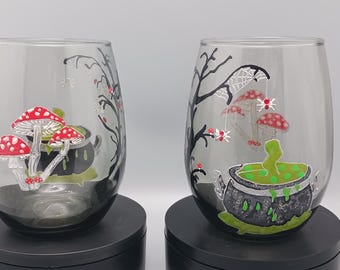 May include: Two stemless wine glasses with Halloween-themed designs. One glass features red and white mushroom illustrations, while the other has a cauldron with green liquid and spiderwebs. The glasses are clear with black and red accents.