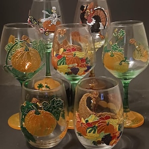 Puede incluir: Una colección de siete copas de vino transparentes y vasos sin pie con diseños pintados a mano con temática otoñal. Las copas presentan ilustraciones de calabazas, calabazas y pavos en tonos naranja, verde y marrón. Los tallos son verdes.