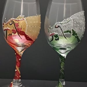May include: Two clear wine glasses featuring hand-painted dragon designs. One dragon is gold and red, the other is silver and green. The stems are decorated with matching colours. A unique addition to any glassware collection.