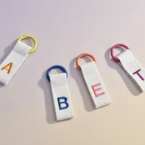 May include: Four white fabric keychains with embroidered letters spelling "A, B, E, T". Each keychain has a different colored ring: yellow, blue, orange, and pink. The letters are embroidered in matching colors.