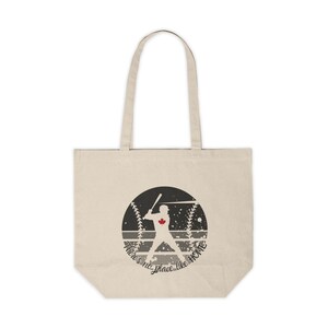 Op de afbeelding: Een beige tote bag met een zwart-witte afbeelding van een honkballer in een cirkel met de tekst "There's no place like home".