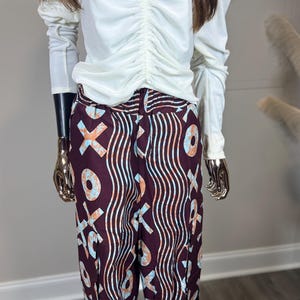 May include: A white long-sleeve top with a ruched front is paired with patterned pants. The pants feature a dark burgundy background with a repeating pattern of wavy lines and X and O shapes in light blue and orange.
