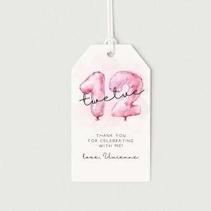 May include: A white gift tag with a pink watercolour background and pink balloon-shaped numbers forming the number 12. The word "twelve" is written in cursive above the numbers. The tag includes the text "Thank you for celebrating with me!" and "love, Vivienne."