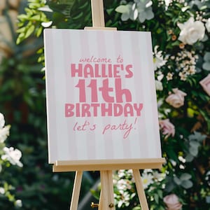 Op de afbeelding: Een rechthoekig bord op een houten ezel met een lichtroze en witte verticaal gestreepte achtergrond. De tekst luidt: welcome to HALLIE'S 11th BIRTHDAY let's party! in vetgedrukte roze letters, met de naam en leeftijd in een groter lettertype.