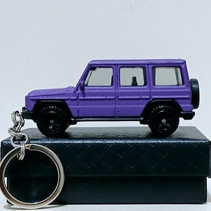 Könnte beinhalten: Ein violetter SUV-Schlüsselanhänger aus Druckguss mit schwarzen Rädern und einem Ersatzreifen. Das Miniaturfahrzeug steht auf einer schwarzen Box, an der ein silberner Schlüsselring befestigt ist. Das Spielzeugauto ist eine detaillierte Nachbildung.