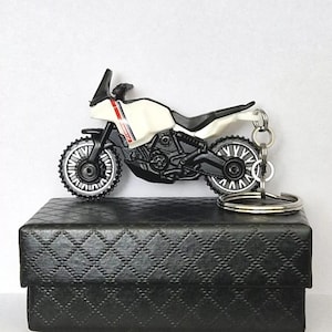 Może przedstawiać: Brelok do kluczy z motocyklem w kolorze białym i czarnym z srebrnym kółkiem. Motocykl znajduje się w czarnym pudełku z diamentowym wzorem.