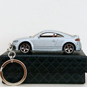 May include: A silver toy car keyring with a red interior is displayed on a black, quilted box. The car features detailed wheels and a spoiler. A large silver keyring is attached.
