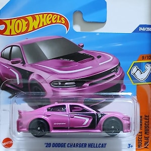 Puede incluir: Un coche de juguete Hot Wheels rosa de un Dodge Charger Hellcat 2020. El coche tiene detalles en negro y ruedas negras. El embalaje incluye el logotipo de Hot Wheels y el texto "Muscle Mania".