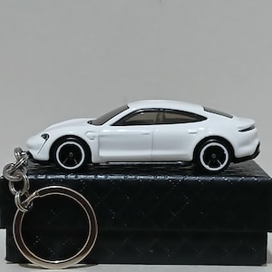 Könnte beinhalten: Ein weißer Spielzeugauto-Schlüsselanhänger mit schwarzen Rädern und einem silbernen Schlüsselring. Das Auto hat ein schlankes, modernes Design und steht auf einer schwarzen, strukturierten Box. Der Schlüsselanhänger ist eine Miniatur-Nachbildung eines Sportwagens.