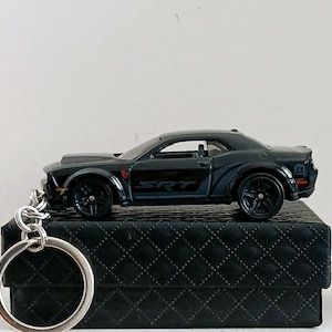 Puede incluir: Llavero negro Dodge SRT Challenger. El coche en miniatura presenta ruedas negras, una carrocería detallada y el logotipo SRT. El llavero incluye un anillo de metal plateado para sujetar las llaves. El coche se exhibe sobre una superficie negra texturizada.