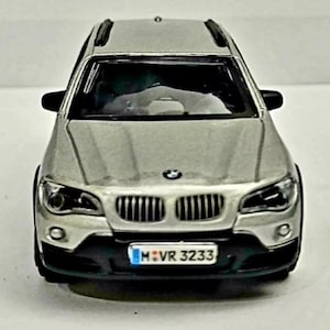 May include: A silver toy car model of a BMW X5 SUV with a black grill and black trim. The car has a license plate that reads "M VR 3233".