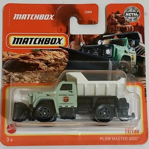 May include: A Matchbox toy truck, the Plow Master 6000, in a green and white color scheme. The toy truck has black wheels and a plow blade. The packaging is orange and features the Matchbox logo and the text "National Parks".