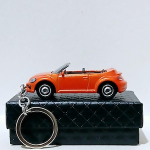Op de afbeelding: Oranje cabrio autosleutelhanger. De miniatuurauto heeft zwarte wielen en een zilveren voorruitframe. De sleutelhanger is bevestigd aan een zilveren metalen ring. De auto staat op een zwarte, getextureerde doos.