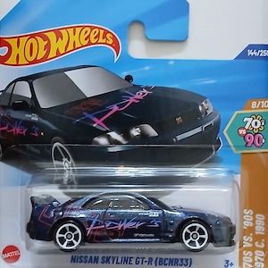 Peut inclure: Modèle réduit Hot Wheels d'une Nissan Skyline GT-R (BCNR33) bleu foncé. La voiture arbore l'inscription "Fower's" en rose et violet, des roues blanches et un aileron arrière. L'emballage comprend le logo Hot Wheels et la marque "70s vs 90s".
