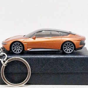May include: A miniature orange sports car keychain with a black roof and gray wheels. The car sits atop a black box, with a silver key ring attached. The car's design is sleek and modern, suggesting a collectible item or a gift for car enthusiasts.