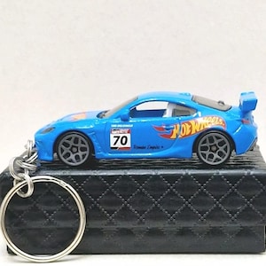 Peut inclure: Une voiture jouet bleue Hot Wheels avec un porte-clés noir. La voiture a une bande blanche et les mots "Hot Wheels" et "Roman Empire" sur le côté. La voiture a le numéro "70" sur le côté.