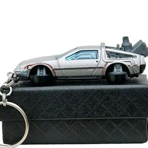 Könnte beinhalten: Ein silberner DeLorean-Zeitmaschinen-Schlüsselanhänger mit einem schwarzen Lederetui. Der Schlüsselanhänger ist eine detaillierte Nachbildung des legendären Autos aus den Filmen Zurück in die Zukunft.