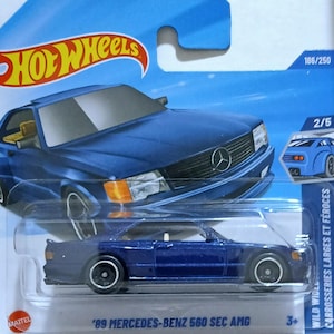 May include: A blue Hot Wheels die-cast toy car of a '89 Mercedes-Benz 560 SEC AMG. The car has black wheels and a silver Mercedes-Benz logo. The packaging includes the Hot Wheels logo and the text "WILD WIDE CARROSSERIES LARGES ET FÉROCES".