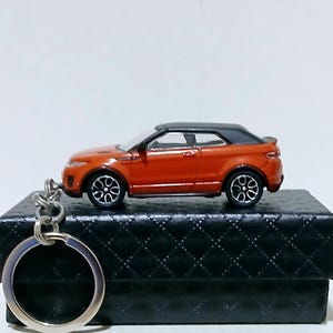 Puede incluir: Llavero de coche de juguete naranja y negro. El coche en miniatura tiene un techo negro y ruedas plateadas. El llavero está unido a un anillo de metal plateado. El coche está sobre una caja negra texturizada.