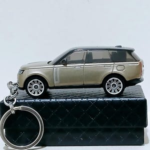 May include: A miniature tan SUV keyring with a black roof and silver wheels. The keyring is attached to a silver key ring. The SUV is sitting on a black, textured box.