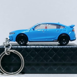 May include: A light blue die-cast car keyring with black wheels and a silver key ring. The miniature car has a spoiler and is resting on a black, textured box. The car is a miniature replica of a sedan.