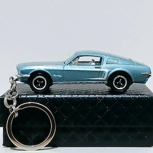 May include: A light blue die-cast model of a classic Mustang car, attached to a silver keychain. The car has black wheels and white accents. The keychain is resting on a black, quilted box. The car is approximately 3 inches long.