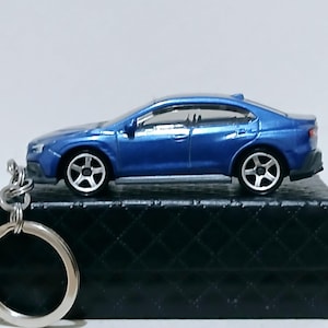 Puede incluir: Un llavero de coche modelo azul. El coche en miniatura tiene un diseño elegante con ruedas negras y un llavero plateado. El coche se exhibe en una caja negra texturizada.