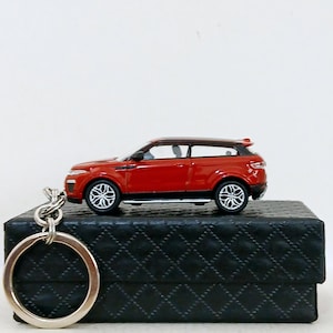 May include: A red miniature SUV keyring with black trim and silver wheels. The keyring is attached to a silver metal ring. The car is resting on a black, textured box.