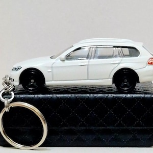 Puede incluir: Llavero de coche modelo de fundición a presión blanco de un BMW familiar. El coche está sobre una caja negra con un patrón acolchado.