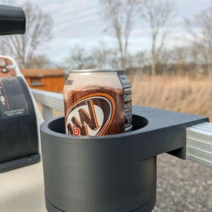 May include: A black, 3D-printed cup holder attached to a metal rail, holding a can of A&W root beer. The can is brown and features the A&W logo. The background shows a field and trees under a cloudy sky.