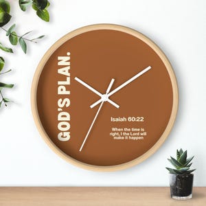 May include: A round wall clock with a light wood frame and a brown face. The clock features white hands and the text "GOD'S PLAN." and "Isaiah 60:22" with a quote. A small succulent plant is in the lower right corner.