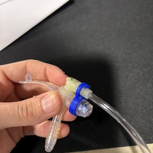 Peut inclure: Un dispositif médical en plastique transparent avec une pince bleue et un tube en plastique transparent. Le dispositif comporte un petit récipient en plastique transparent sur lequel est imprimé le texte "BAX 6 ML".