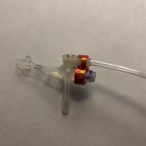 May include: A medical device with clear plastic tubing and connectors. The device has a central hub with red and orange bands, and a small blue component. The tubing extends to the right, and a small clear extension is on the left.
