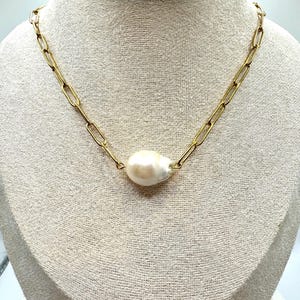 May include: A gold-colored chain necklace with a large, off-white baroque pearl pendant. The chain has a paperclip link design. The necklace is displayed on a neutral-colored mannequin.