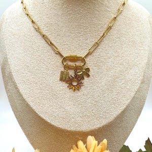 Puede incluir: Collar dorado con un diseño de cadena de clip. El collar presenta un cierre con tres colgantes: la letra "M", un sol y un trébol de cuatro hojas. El collar se exhibe en un maniquí.