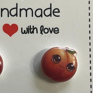 Pode incluir: Um par de brincos de botão artesanais em forma de pêssego. Cada brinco apresenta um design de pêssego sorridente com olhos grandes e uma pequena folha verde. O cartão diz "Handmade with love."