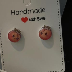Pode incluir: Um par de brincos de tarracha com um design de pêssego de desenho animado e uma pequena coroa de flores no topo. Os brincos estão expostos num cartão branco com o texto Handmade with love e um ícone de coração vermelho impresso.
