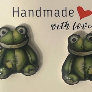 May include: Two green frog-shaped handmade decorations with smiling faces and yellow patches. The frogs are on a white background with the words "Handmade with love" and a red heart.