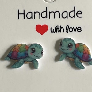 Könnte beinhalten: Ein Paar handgefertigte Schildkrötenohrringe. Die Schildkröten sind türkis mit bunten Puzzleteilmuster auf ihren Panzer. Die Ohrringe befinden sich auf einer weißen Karte mit den Worten "Handmade with love" in Schwarz und Rot gedruckt.