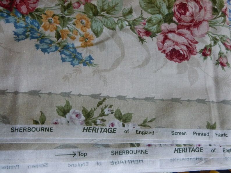 Puede incluir: Primer plano de una tela beige con un patr&oacute;n floral con rosas rosas, flores amarillas y campanillas. La tela tiene un borde blanco con el texto "SHERBOURNE HERITAGE of England Screen Printed Fabric" repetido.
