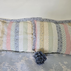 May include: Two decorative throw pillows with a patchwork design featuring stripes and floral patterns in shades of pink, beige, and blue. The pillows are on a light blue bedspread with a floral pattern.