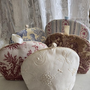 May include: Four different tea cozy covers. One is white with embroidered flowers and swirls. One is red with a toile pattern. One is brown with a toile pattern. One is blue and white striped with a floral pattern.