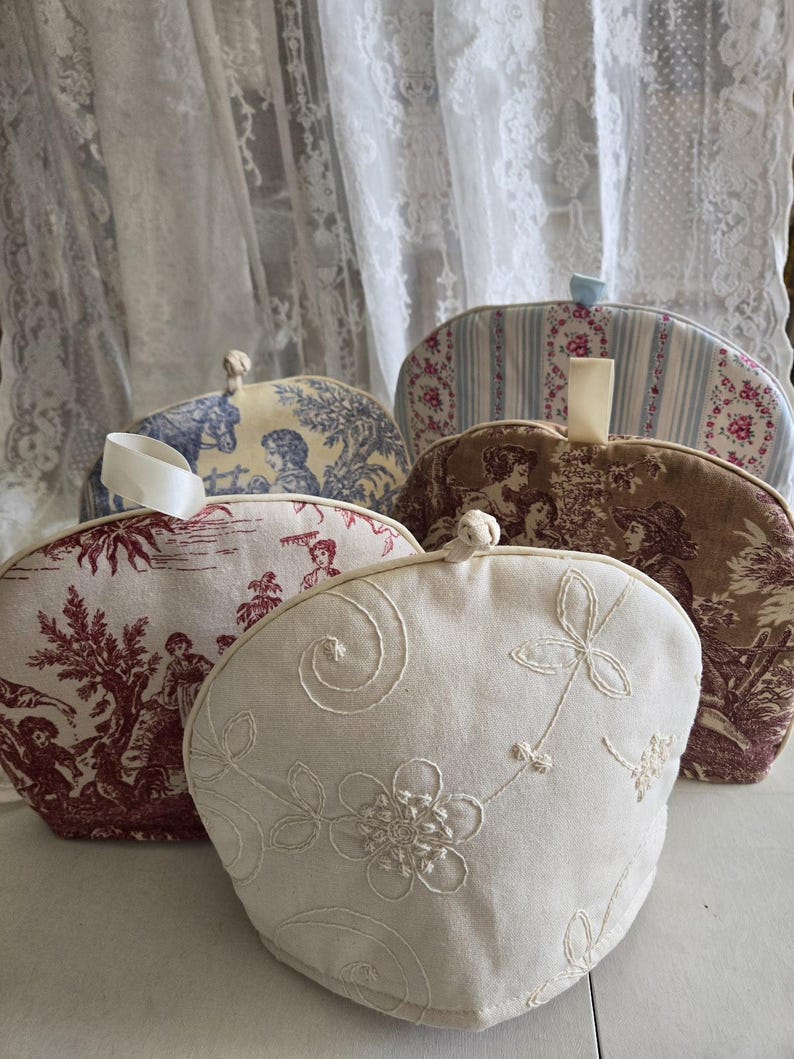 May include: Four different tea cozy covers. One is white with embroidered flowers and swirls. One is red with a toile pattern. One is brown with a toile pattern. One is blue and white striped with a floral pattern.
