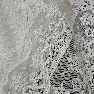 Peut inclure: Gros plan sur un tissu en dentelle blanc cassé avec des motifs floraux et géométriques complexes. La dentelle présente des détails délicats et un fond en résille fine, créant un aspect texturé et élégant. L'image met en évidence le savoir-faire de la dentelle.