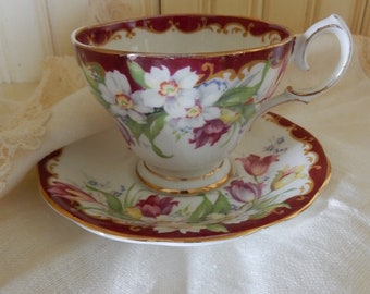 May include: A vintage teacup and saucer set with a floral design. The cup and saucer are white with a red rim and gold accents. The floral design features white and pink flowers with green leaves.